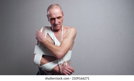 Injured Senior Man Tied Hard With White Bandage On Bare Torso Examines Broken Forearm Standing At Room Grey Wall At Home Close View.