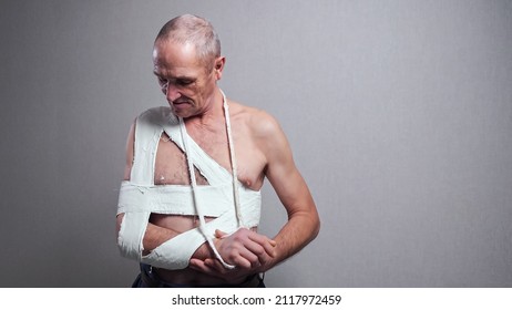 Injured Senior Man Tied Hard With White Bandage On Bare Torso Examines Broken Forearm Standing At Room Grey Wall At Home Close View.