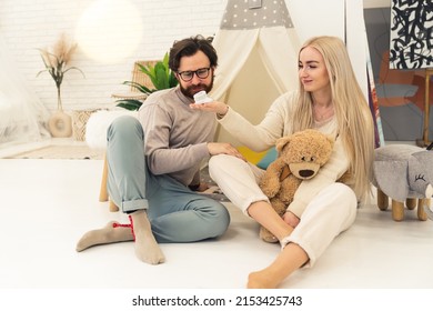 Inner shot of a caucasian excited couple in their 20s sitting on the floor in a nursery with their legs crossed. Woman holding a small baby boot and a giant teddy bear enjoying future motherhood. High