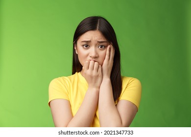 Innocent insecure timid asian scared girl panicking, standing afraid victim terrified, touch cheek shocked, frowning stunned, biting fingernails, anxiously stare camera, green background
