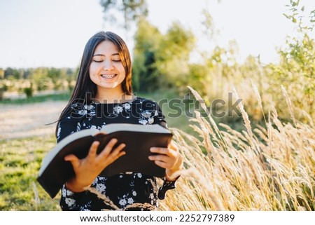 Innocent young religious girl with braces studying the bible, outside in the field at sunset. Spiritual revival