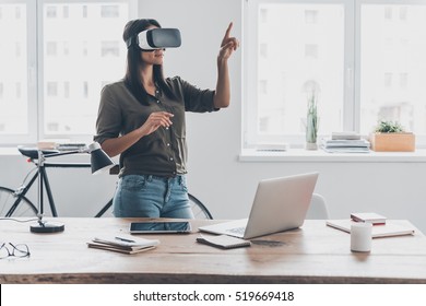 Innovation in business. Confident young woman in virtual reality headset pointing in the air while standing near her working place in office