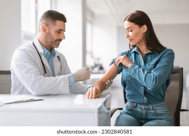 Inoculation time. Doctor man wearing gloves and making vaccination shot to young female patient, inserting syringe to her arm, woman getting vaccine shot against viruses in modern clinic - Powered by Shutterstock