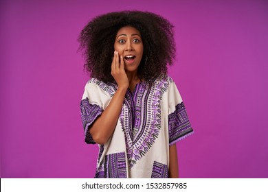 Inoor Photo Of Amazed Young Dark Skinned Woman With Curly Hair Keeping Raised Palm On Her Face And Looking At Camera With Suprised Face, Isolated Over Purple Background