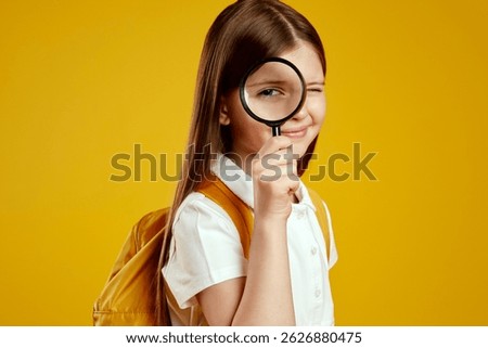 Inquisitive child with a backpack peeks through a magnifying glass, displaying a playful and curious demeanor against a yellow classroom background. Ideal for educational content and school promotions