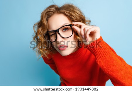 Inquisitive female in red sweater adjusting glasses and looking at camera closely against blue background