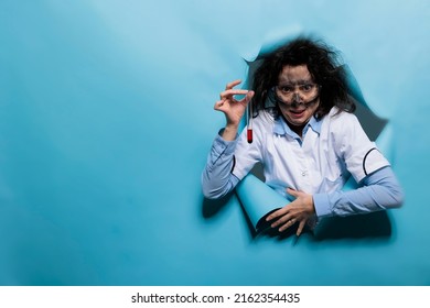 Insane biochemistry specialist with messy hair and dirty face grinning creepy while having glass test tube filled with chemical mix. Crazy chemist with wacky goofy look on blue background. Studio shot