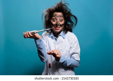 Insane lab worker grinning creepy while dripping toxic compound from pipette into glass test tube while looking at camera on blue background. Crazy scientist with messy hair and dirty face using pipet