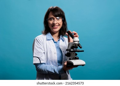 Insane looking scientist smiling creepy while having modern microscope instrument on blue background. Lunatic chemist with gruesome smile having electronic magnifying laboratory instrument.