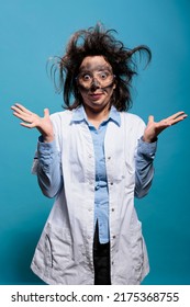 Insane And Silly Looking Crazy Scientist With Messy Hairstyle And Dirty Face Does Not Understand Where Experiment Went Wrong. Mad Chemist With Goofy Expression Standing On Blue Background. Studio Shot