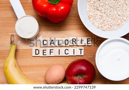 Inscription calorie deficit next to fruit, eggs, protein powder and oatmeal. Weight reduction. Healthy sources of proteins and carbohydrates