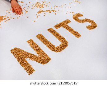 The inscription "NUTS" from the peeled kernels of cedar cones on a white table, a child's pen takes nuts. Nuts, vegetable protein, vegetarianism, healthy snack, proper nutrition. Healthy lifestyle