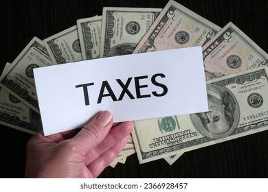 the inscription on the paper taxes lies on top of a large pile of dollar USA money on a dark wooden table, top view, concept - Shutterstock ID 2366928457