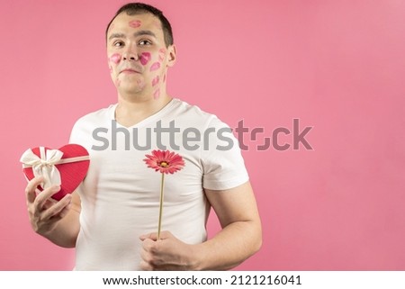 An insecure and in love man with a flower and a gift for Valentine's Day, confused by the kisses of a woman. A surprised, confused man with traces of kisses on his face.