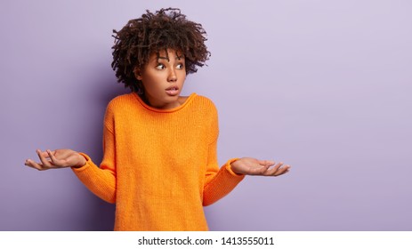 Insecure puzzled dark skinned female has Afro hairstyle, spreads hands with doubt, dressed in orange jumper, isolated over purple background, blank copy space. Woman faces dilemma, being unsure
