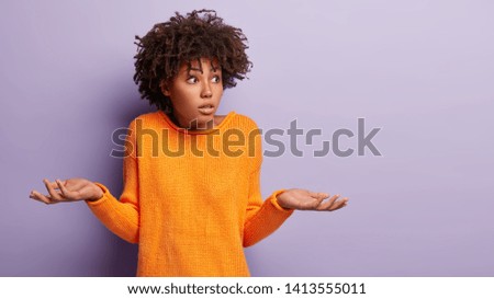 Insecure puzzled dark skinned female has Afro hairstyle, spreads hands with doubt, dressed in orange jumper, isolated over purple background, blank copy space. Woman faces dilemma, being unsure
