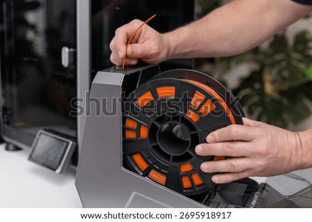 Inserting an orange filament spool into a 3D printer holder, concept of preparing printing material such as PLA, ABS, PETG, or TPU for additive manufacturing.