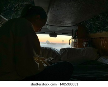 The Inside Of A Backpackers Camper Van Used To Travel Around Australia. A Surfboard Hangs On The Roof, A Female Traveler Reads On Her Phone As The Sun Rises Over The Ocean At Currumbin, Gold Coast