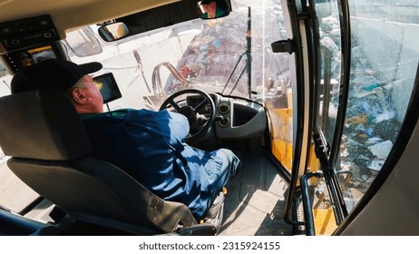 Inside cab view of a man driving heavy machine vehicle, a front loader grabbing garbage and loading a truck, fast motion. Waste management concept.