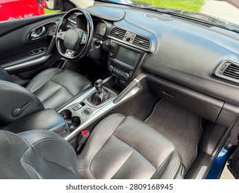 Inside a car, there are two front seats, a dashboard with controls, and a gear stick. The setting is bright and the vehicle is parked.