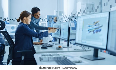 Inside Factory Office: Male Project Supervisor Talks to a Female Industrial Engineer who Works on Computer, Talk. In Workshop: Professional Workers Use High-Tech Industry 4 CNC Machinery, Robot Arm.
