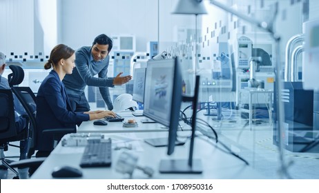 Inside Factory Office: Male Project Supervisor Talks To A Female Industrial Engineer Who Works On Computer, Talk. In Workshop: Professional Workers Use High-Tech Industry 4 CNC Machinery, Robot Arm.