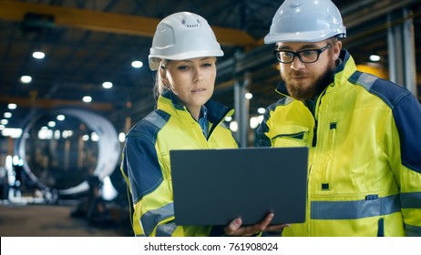 Inside The Heavy Industrial Factory Female Industrial Engineer Holds Laptop And Has Discussion With Project Manager. They Wear Hard Hats And Safety Jackets. In The Background Metalwork In Progress.