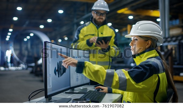 Inside the Heavy Industry, Factory Female\
Industrial Engineer Works on Personal Computer She Designs 3D\
Engine Model, Her Male Colleague Talks with Her and Uses Tablet\
Computer. Low Angle\
Shot.