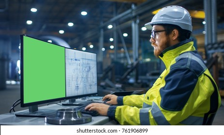 Inside The Heavy Industry Factory Industrial Engineer Works On The Personal Computer With Two Screen. One Is Green Mock-up Screen And Other With Blueprints.