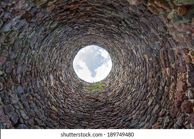 The inside of an old industrial chimney shaft photographed from the bottom - circular stone wall with tree growing from it and blue sky with white clouds in the opening in the centre, horizontal - Shutterstock ID 189749480
