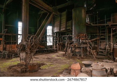 Inside an old industrial forge workshop, rusted machinery stands among scattered tools. Sunlight filters through dirty windows, casting greenish hue on the dusty floor, creating a nostalgic atmosphere