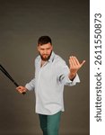 Inside a studio, a confident man with a beard stands with a baseball bat in hand. He gestures toward the viewer, radiating an artistic vibe, surrounded by an abstract backdrop.