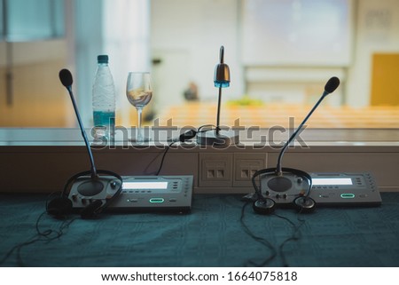 Inside of translation or  interpreting booth. Desk with microphones and audio board, with visible conference hall in the background.