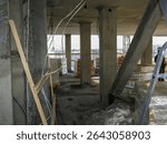 Inside view of a building under construction shows concrete pillars, support beams, and construction materials.