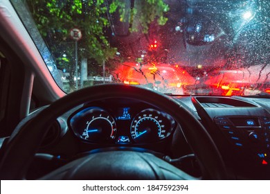 Inside view of a car, from the driver's point of view, stopped in traffic light on a rainy day. Dashboard lights, raindrop windshield and exterior lights.
