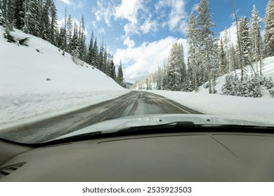 Inside view of a car driving through snow covered mountains in a forest with blue sky in the background