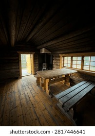 Inside view of a rustic log cabin in Sweden with wooden tables, benches, a stove, and an open doorway overlooking a forest.