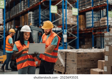 Inside a warehouse, a focused african american woman and a caucasian man wearing safety gear are reviewing laptop data among shelves, portraying organizational skills and a results-driven mindset. - Powered by Shutterstock