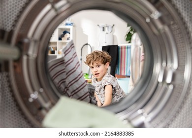 Inside The Washing Machine, A Little Boy Is Putting Colorful Clothes Into The Drum Through The Open Door, Helping Father With Household Chores.