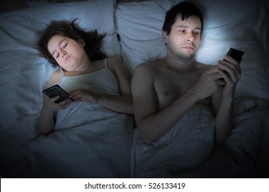 Insomnia Concept. Young Couple Is Lying In Bed And Cant Sleep. They Are Browsing Internet Or Texting With Smartphone.