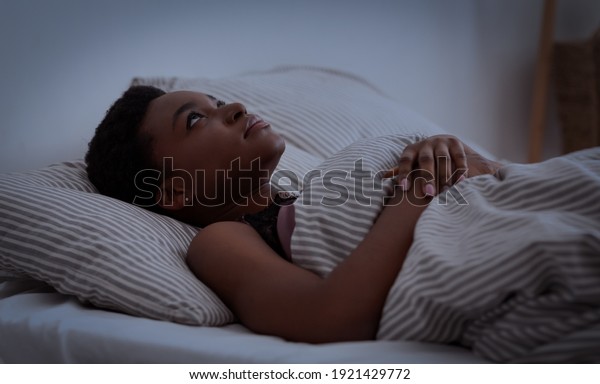 Insomnia, sleep problems, health care and bed
sleep. Depressed young african american young woman lies on bed
with blanket with open eyes, in dark bedroom interior, copy space,
profile, panorama