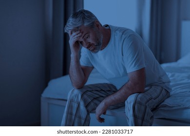 Insomnia, sleeping disorder concept. Sleepless unhappy grey-haired middle aged man wearing pajamas sitting on bed at home, touching his head, cant sleep at night, copy space - Shutterstock ID 2281033717