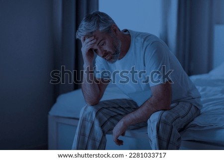 Insomnia, sleeping disorder concept. Sleepless unhappy grey-haired middle aged man wearing pajamas sitting on bed at home, touching his head, cant sleep at night, copy space