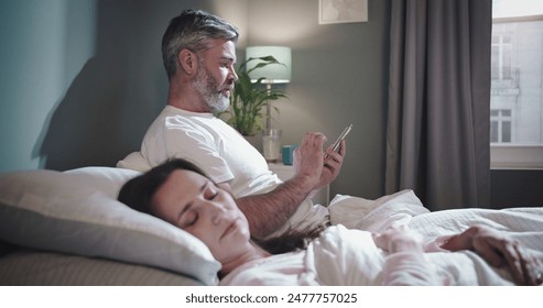 Insomnia. Waist up portrait view of the caucasian man laying at the bed near his wife and using his smartphone while his woman sleeping - Powered by Shutterstock