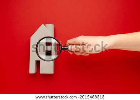 Inspection of real estate. Miniature house and hand with a magnifying glass on a red background. Search for assets.