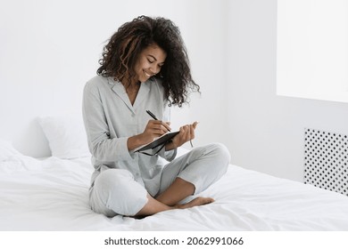 Inspiration, Imagination Concept. Smiling Young Afro American Woman Sitting On Bed In Nightwear, Writing In Personal Journal. Happy Student Makes Notes In Diary Spending Morning At Home