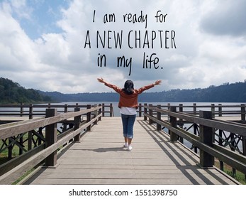 Inspirational motivational quote - I am ready for a new chapter in my life. With young woman standing on wooden bridge, hands raised with open arms against the blue sky & lake view background.