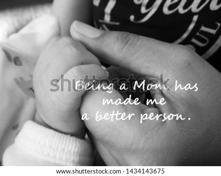 Inspirational quote- Being a Mom has made me a better person. With blurry image of a fragile little baby new born hand and fingers holds by her his mother hand in black and white.