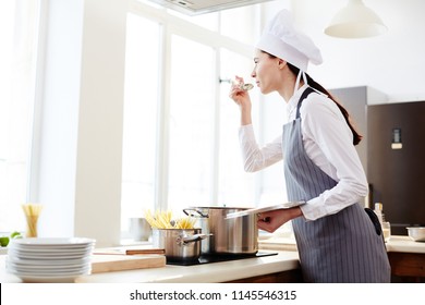 Inspired attractive talented female pasta maker in hat and apron standing at counter with induction cooker and tasking soup while cooking in restaurant kitchen - Powered by Shutterstock