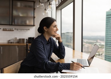 Inspired attractive young 30s Indian woman sit at table in domestic or office kitchen distracted from work on laptop looks out window and daydreams, planning, having business vision, dreaming concept